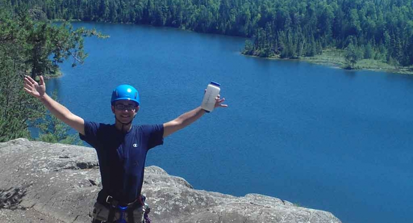 a person wearing rock climbing gear stands on a cliff above blue water and raises their hands in celebration